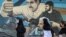 Beberapa perempuan berjalan melewati mural anti-Israel yang menggambarkan pejuang dengan senapan serbu di sepanjang dinding di Alun-Alun Palestina di Tehran pada 14 April 2024. (Foto: AFP)